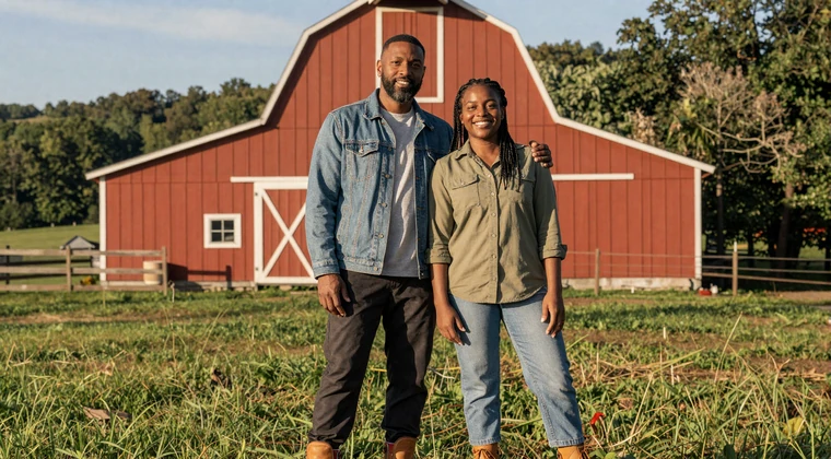 African American couple standing proudly in front of a red barn on their North Carolina farm, Black farmers dating success story